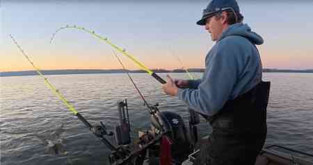 An angler battles a catfish on a neon Hellcat rod at twilight, with the fish’s swirl visible by the boat.