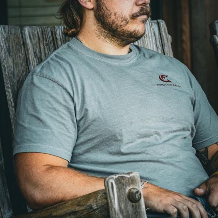 Catch The Fever® Marketing Director Dillon Weaver wearing a grey Catfish Short Sleeve Lifestyle T‑shirt with a small maroon logo, seated on a rustic chair.