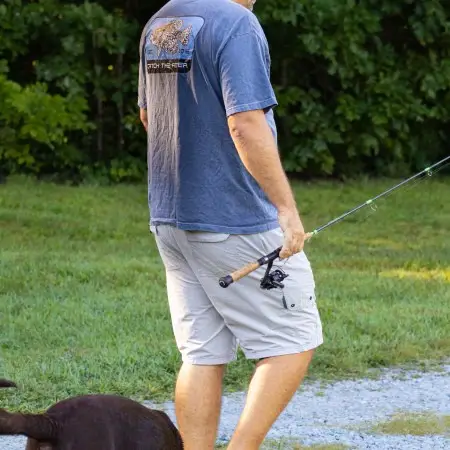 Rear view of a man walking outdoors wearing a dark gray Crappie short sleeve lifestyle T shirt with a large yellow crappie fish graphic and Catch The Fever® logo, holding a fishing rod and accompanied by a brown dog.